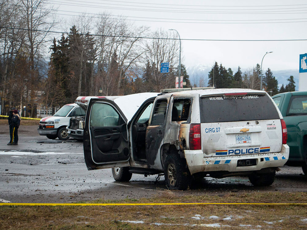 Caution tape blocks off a burned-up RCMP SUV that has its door open. A couple people in uniforms stand in the parking lot beyond the SUV.