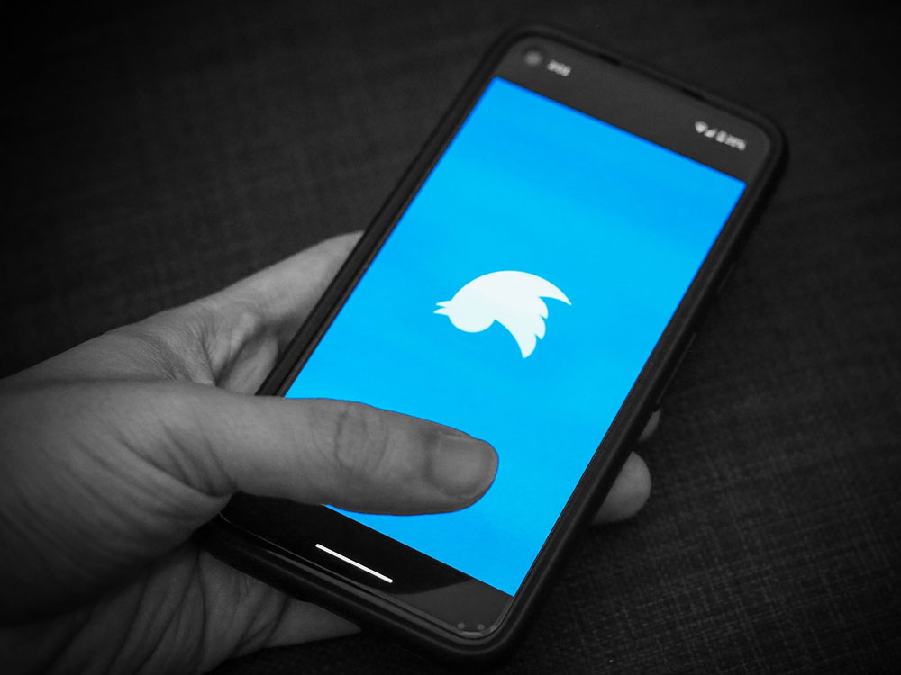 A closeup of a hand, in black and white, holding a phone. It&rsquo;s screen shows a blue field and an upside down Twitter logo. 