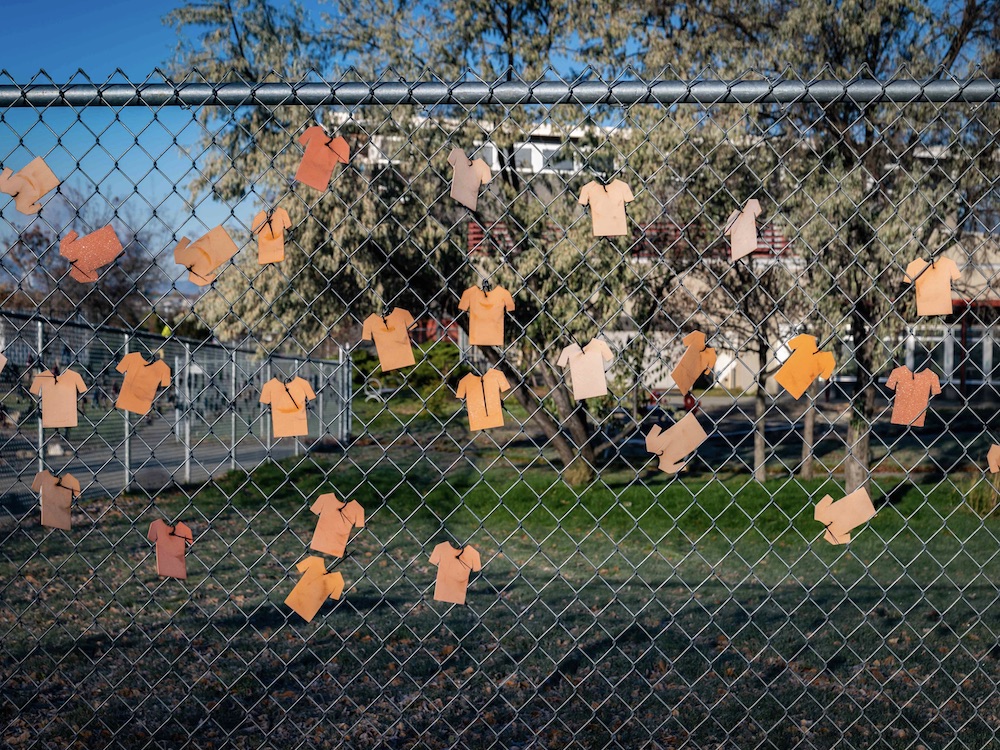 Numerous paper orange shirts are arranged on a chain link fence in front of a public school in Kamloops, B.C. in October 2021. The school is visible in the background behind a stand of trees and a green field. It&rsquo;s a sunny autumn day and the sky is blue.
