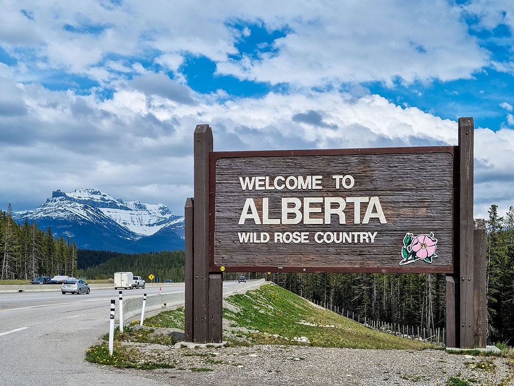 A highway sign says &lsquo;Welcome to Alberta &mdash; Wild Rose Country.&rsquo; Coniferous trees line the four-lane highway. A snow-covered mountain rises against a background of blue skies and light clouds.