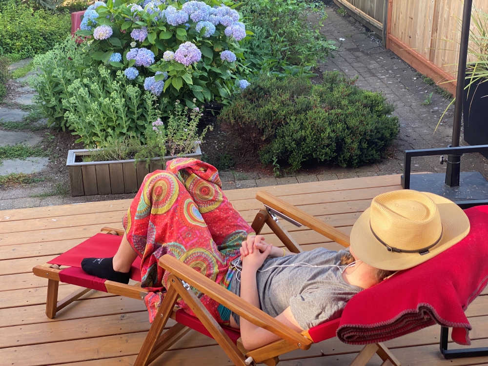  A young woman lies on a red upholstered lawn chair in a garden. Blue hydrangeas blossom across the top of the frame. The woman is wearing a white T-shirt, colourful red pants and black sport socks. A straw hat is covering her face.