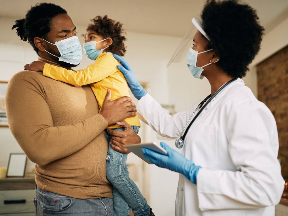 A physician in a white lab coat with a stethoscope around her neck stands to the right of the frame, placing her hand gently on the back of a young child in a yellow shirt to the left of the frame. The child is in their father&rsquo;s arms; he is wearing a beige sweater and jeans. All three are in blue medical masks; the physician has a face shield.