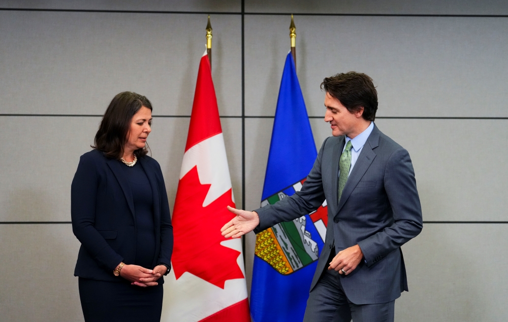 Danielle Smith, in a blue suit, stands with her hands folded at her waist as Justin Trudeau reaches out to shake her hand. He&rsquo;s smiling. Smith is looking at his hand like it&rsquo;s a dead fish. They stand in front of Canadian and Alberta flags.