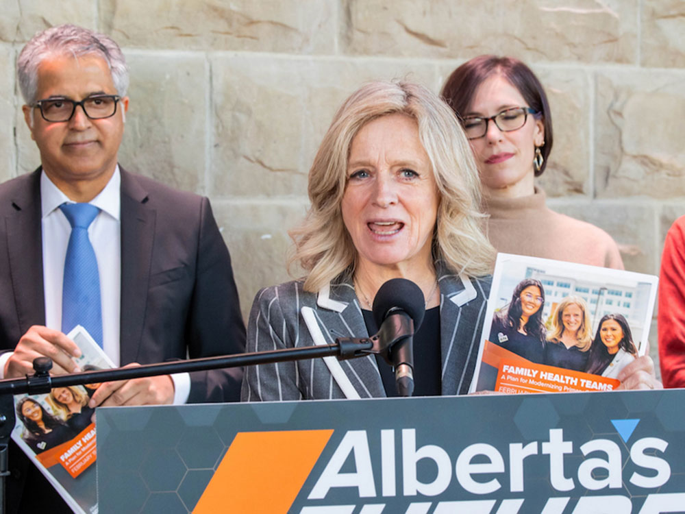 A blonde woman wearing a black shirt and grey striped jacket stands at a podium. She holds a booklet that says &lsquo;Family Health Teams.'