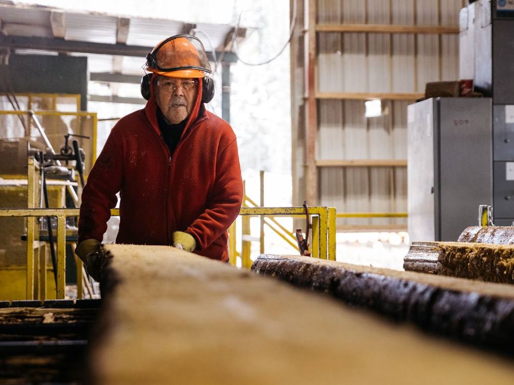 A man wearing a red jacket, a helmet, and ear protection uses both his hands to flip a long log. 