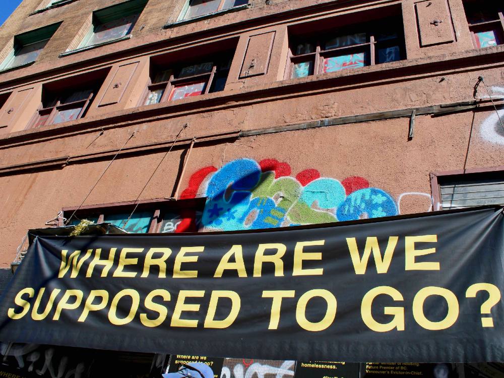 A horizontal black banner with yellow san-serif text reads &ldquo;Where are we supposed to go?&rdquo; in capital letters. The banner hangs across two doorways of a tall brown multi-storey building in the Downtown Eastside. Colourful graffiti can be seen behind the sign. It is a sunny day.