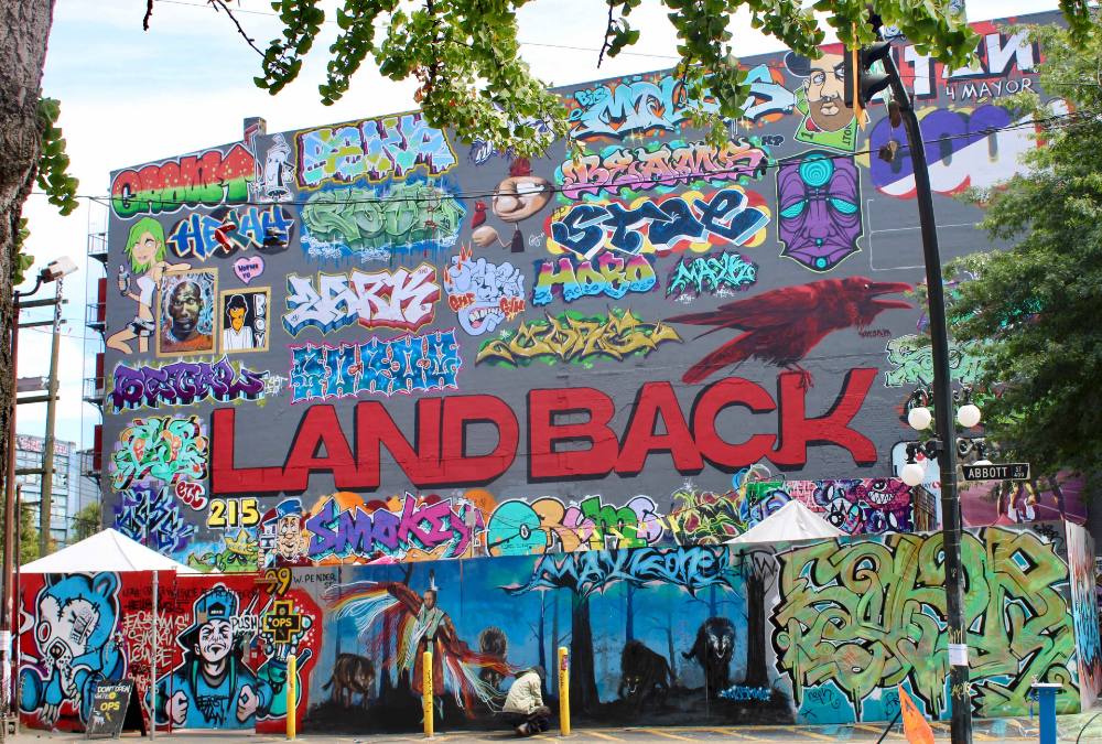 A colourful mural on a dark grey painted wall features the phrase 'Land Back' in red capital letters. It is framed by numerous pieces of colourful artwork by different graffiti artists.