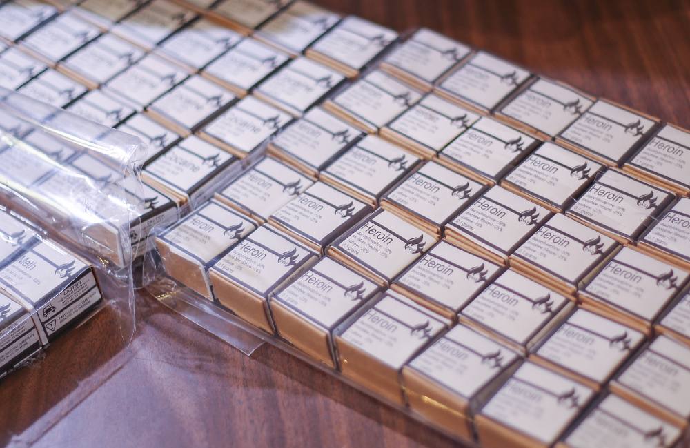 Numerous matchbox-sized boxes with white labels that read &ldquo;Heroin&rdquo; in san serif text line a wooden table. They are arranged in rows in large clear plastic bags with zip closures.