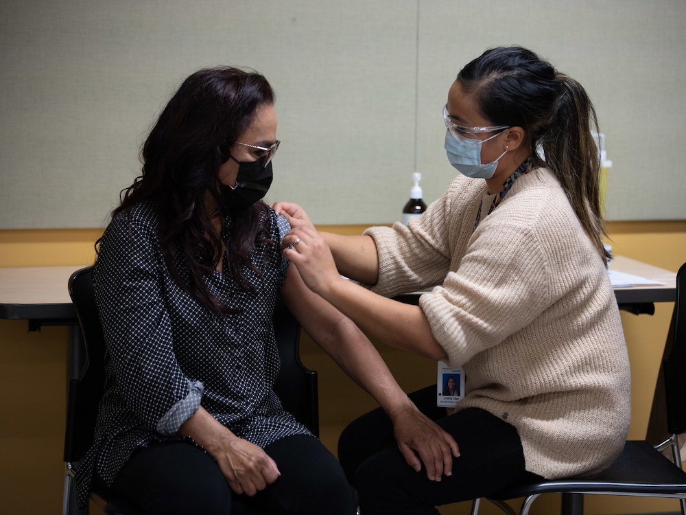 A health-care provider in an off-white sweater applies a bandage to the arm of an Asian woman in a button-down polka-dotted shirt. They are both wearing medical masks and glasses.