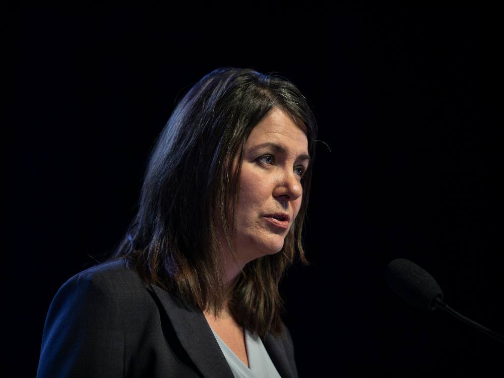 Wearing a black blazer and light-blue shirt, Danielle Smith speaks in front of a microphone.