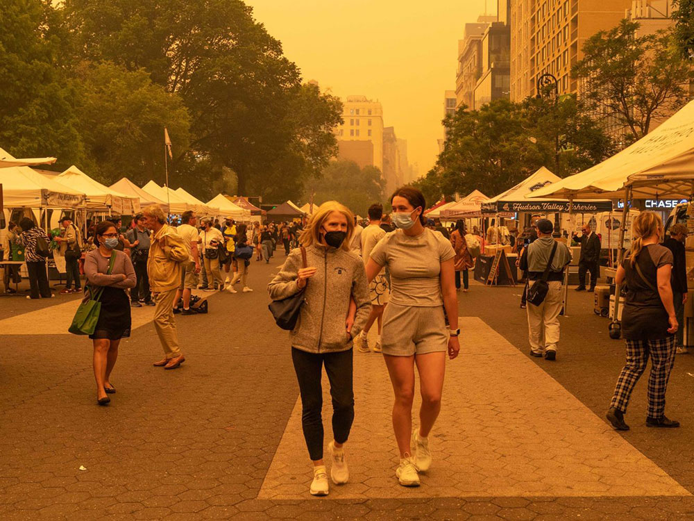 Two women in grey and black athletic apparel walk down the middle of the frame on a tiled concrete walkway. They are both wearing medical masks, as are other patrons amidst the farmers&rsquo; market stalls on both sides of them. The photo has a hazy orange hue from wildfire smoke.