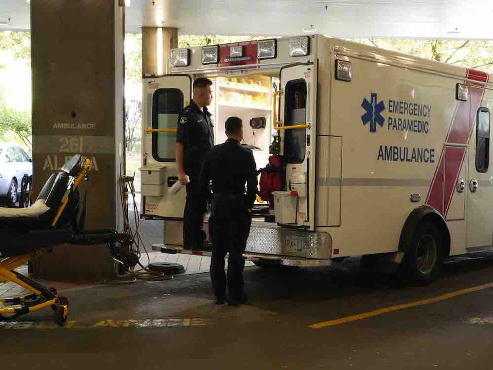 Two Asian men in navy uniforms stand inside the lit back doorway of a B.C. ambulance in a covered parkade. The sun is bright before them. To the left is an empty stretcher.