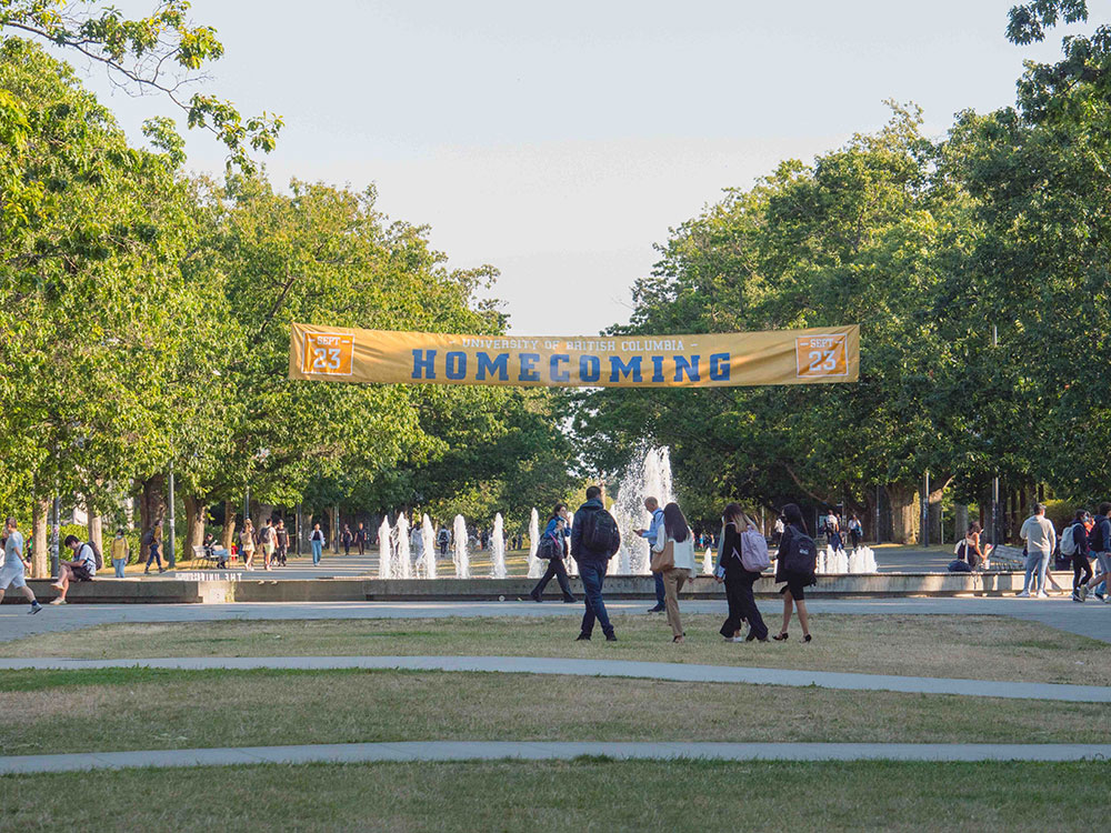 Young adults walk across a tree-lined plaza with a water fountain in the middle. Their backs are turned to the camera and they are wearing backpacks. A yellow banner with the word &ldquo;Homecoming&rdquo; in blue varsity typeface hangs over the fountain between the trees.