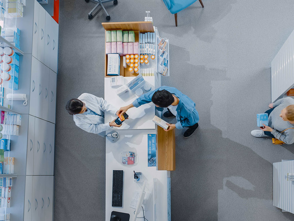 An aerial view of a check-out lineup at a pharmacy with white shelves and light grey flooring. On the left, a pharmacist is helping a customer at a white front counter. Two people are standing in line behind the customer at the counter between white shelves. The person at the back of the line is reaching into one of the shelves.