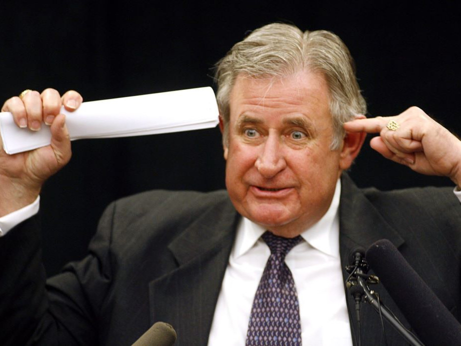 A man in a suit and tie points to his left temple with one hand and to his right temple with a rolled up bunch of papers.