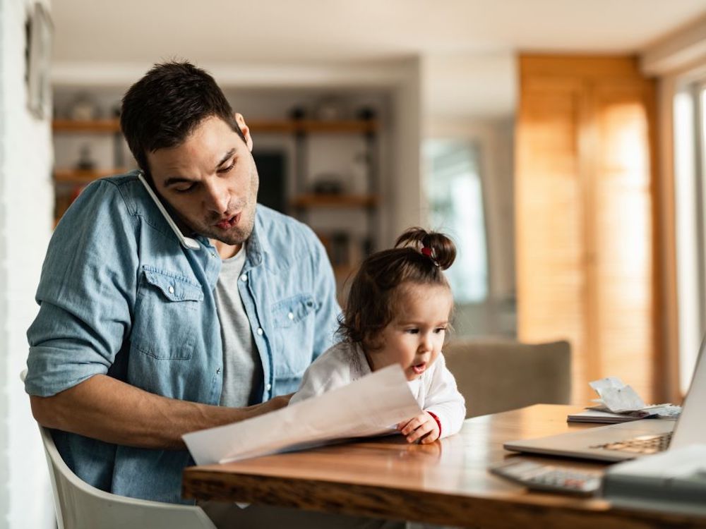 A dark haired man with a toddler daughter on his lap looks over paperwork with a laptop computer open.