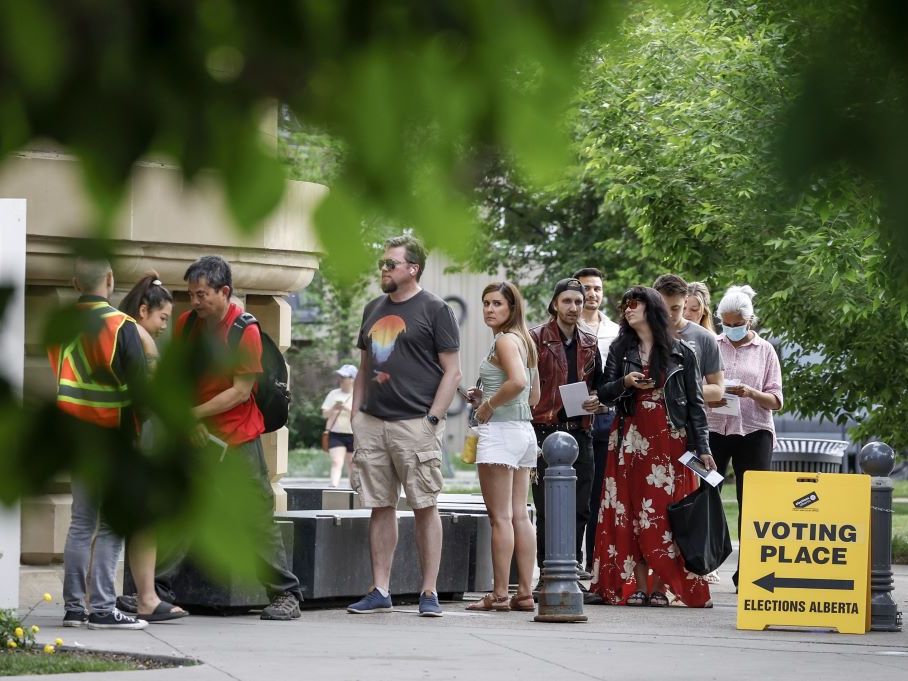 People stand in a line outside near a sign that says, "Voting Place Elections Alberta."