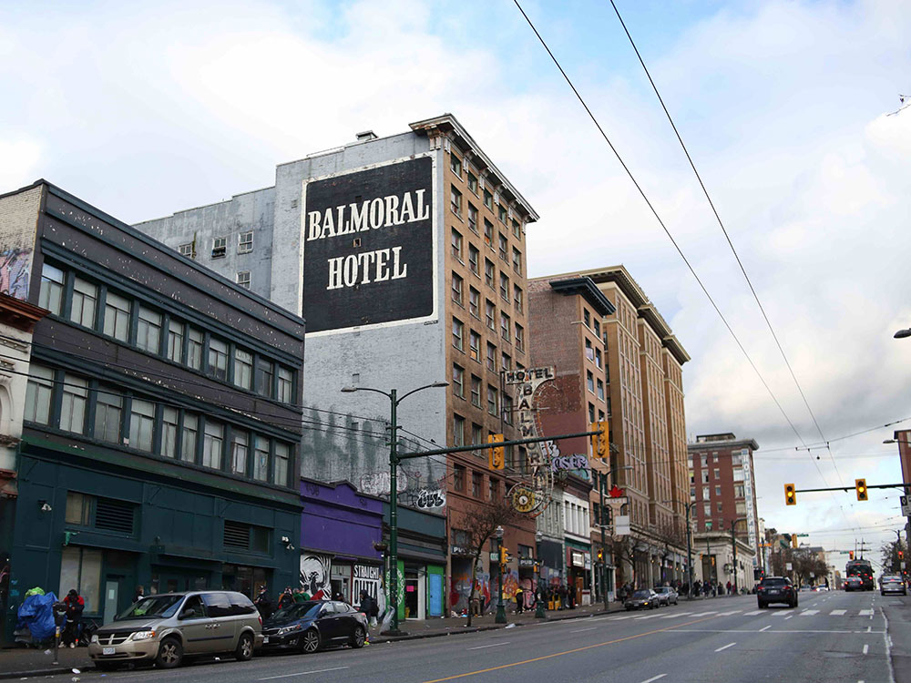 The view of the northeast corner of East Hastings Street at Main in Vancouver. Fluffy white clouds obscure a cold blue sky. A tall brown brick building is painted grey on one side with the words &lsquo;Balmoral Hotel&rsquo; in white all-caps serif script against a black squarish background. It is surrounded by other brick buildings of varying height. A few vehicles are visible parked and on the road. The sidewalk reveals some signs of street activity, but the people are out of focus.