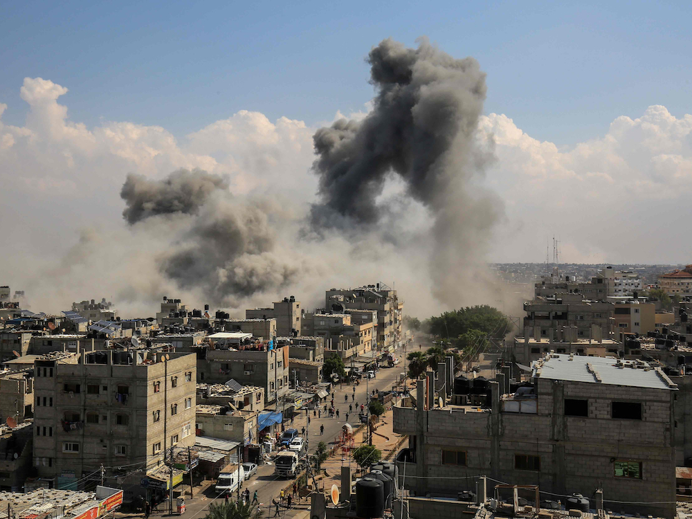 Thick grey plumes of smoke rise above streets of light brown low-rise concrete buildings. The sky behind the smoke is blue with white clouds.