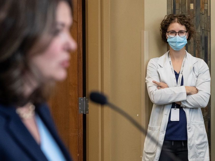 Brown-haired Premier Danielle Smith&rsquo;s face is seen from the side and out of focus. Standing against a wall looking on, and in focus, is a woman with glasses and curly hair, wearing a medical mask, with arms folded and her middle finger extended in a non-demonstrative way.