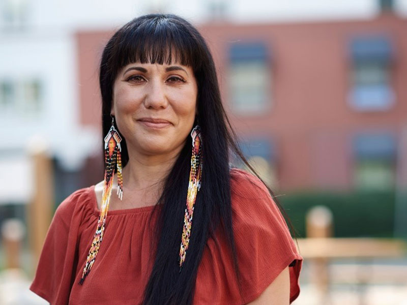 A dark-haired woman wearing long earrings made of beads and feathers smiles slightly.
