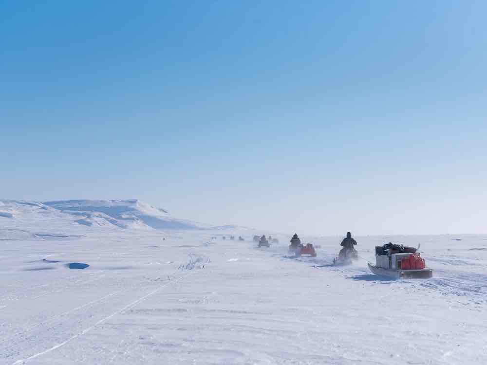 A line of snowmobiles and qamutiks travel away from the camera across a wide, flat expanse of Arctic snow under a big blue sky.