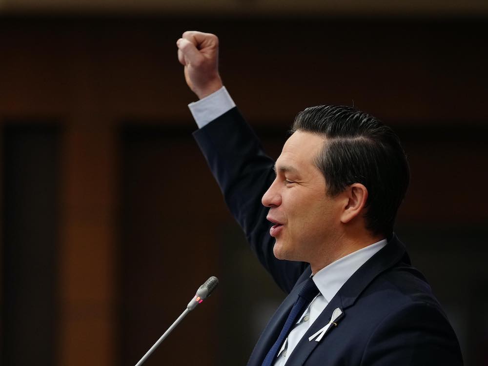 A side shot of a middle-aged white man with greased back black hair wearing a dark blue suit, white shirt and blue tie. He is standing at a podium with one fist raised in the air.