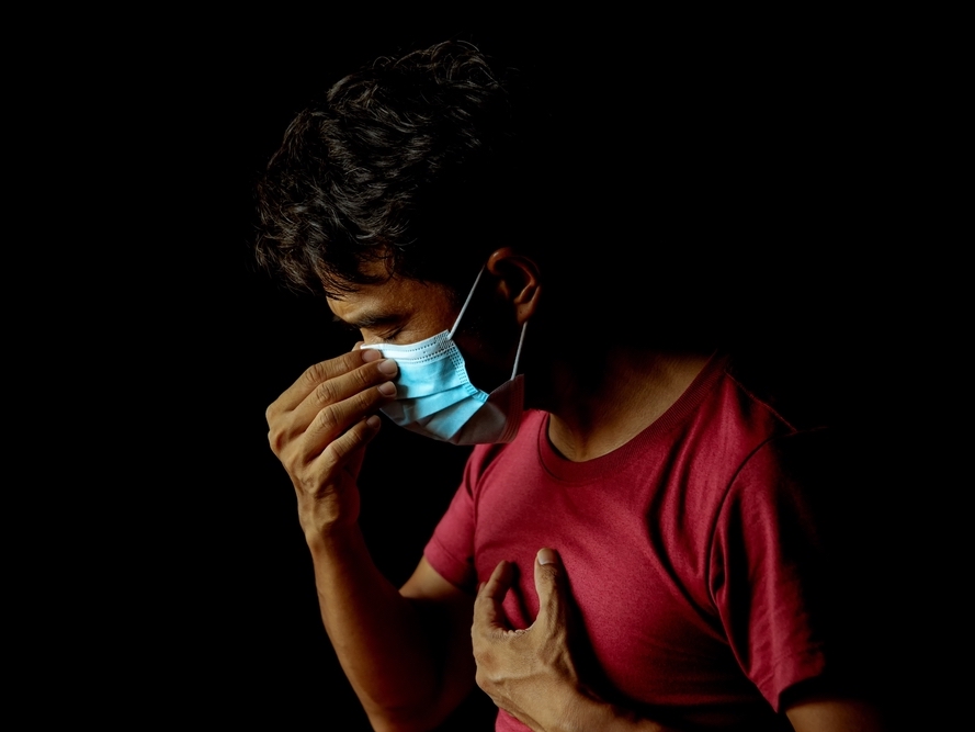 A young adult with short dark hair and wearing a blue mask and red T-shirt has one hand to their face and another on their chest, with eyes closed. The background is black.