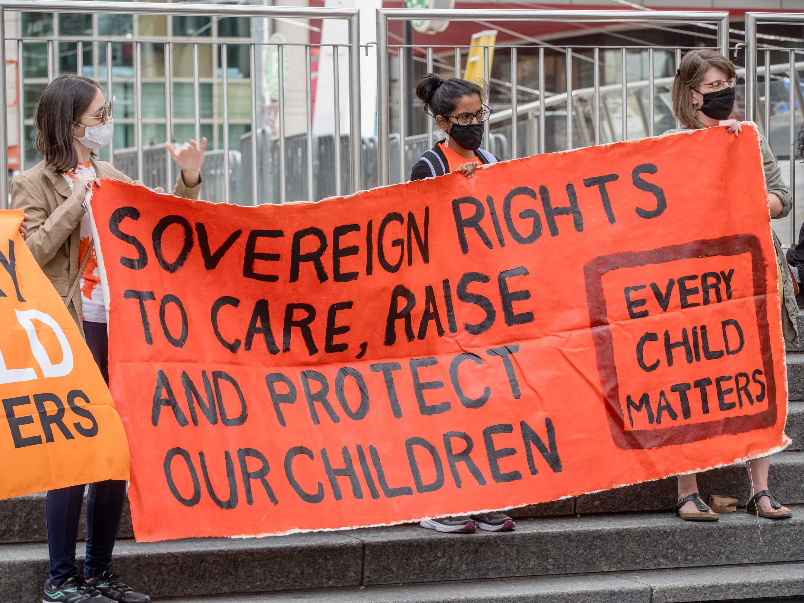 Three people stand on outdoor steps with a sign that says “Sovereign right to care, raise and protect our children” and “Every child matters.”