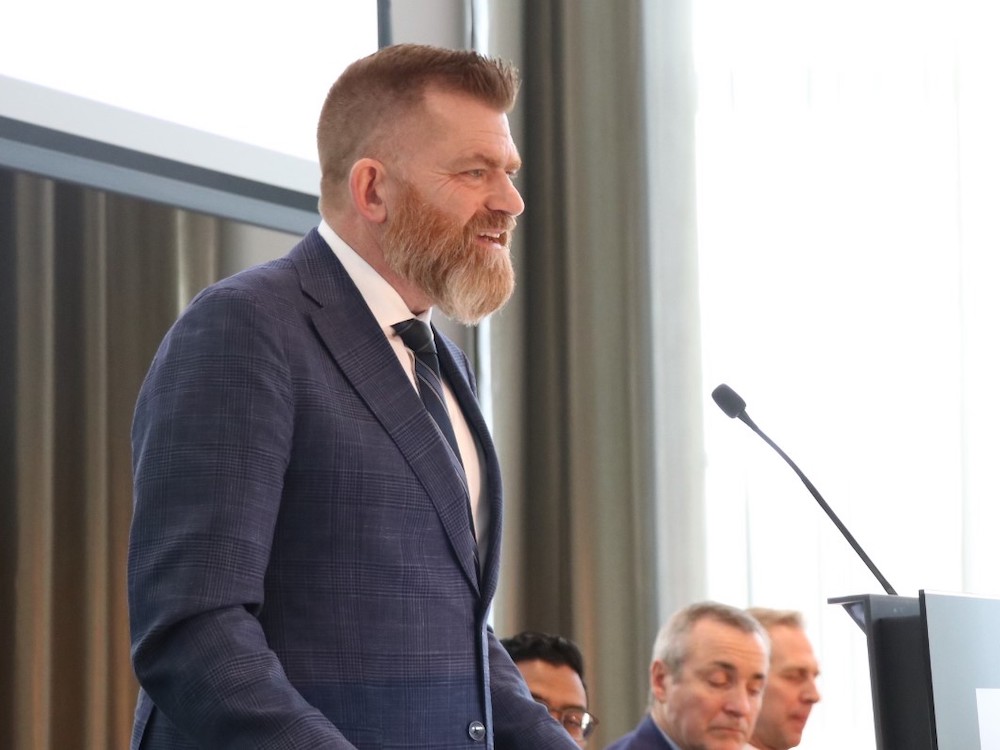 A middle-aged white man with short reddish hair and a full beard stands at a podium with a microphone. He is wearing a blue suit, white shirt and blue striped tie.