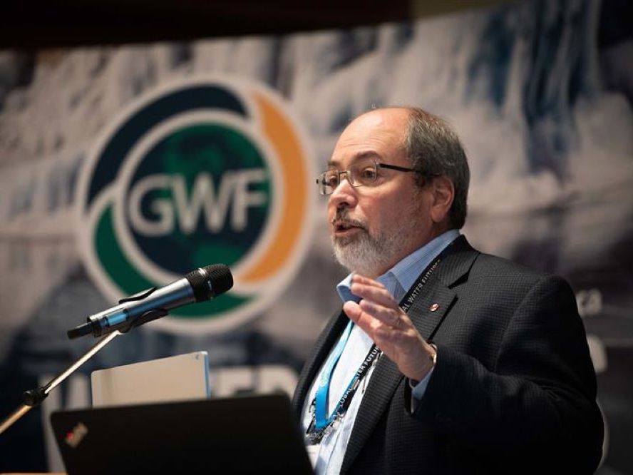 A man with light skin tone, a white-grey beard and greying hair speaks from a podium. He is wearing a suit and a couple of lanyards.