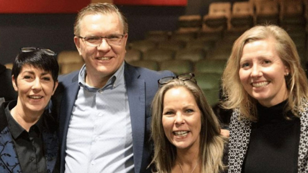 Four white people — from left, a woman with short dark hair, a man with light hair and glasses, and two blond women — pose in a group, smiling.