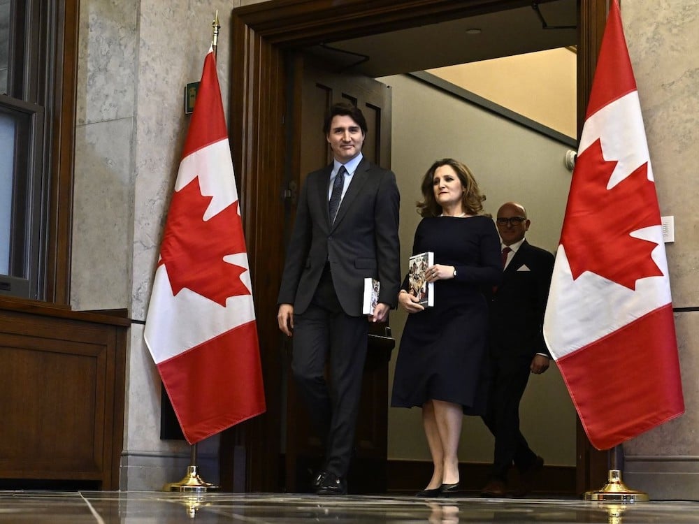 Justin Trudeau, a white man with dark hair wearing a charcoal suit, and Chrystia Freeland, a white woman with shoulder-length brown hair wearing a black dress, emerge from a doorway flanked by two Canadian flags. Trudeau is looking into the camera with a small smile. Freeland is looking ahead.