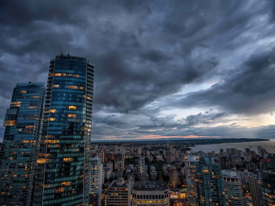 An aerial view shows Vancouver at dusk. Two tall apartment buildings rise toward dark clouds.
