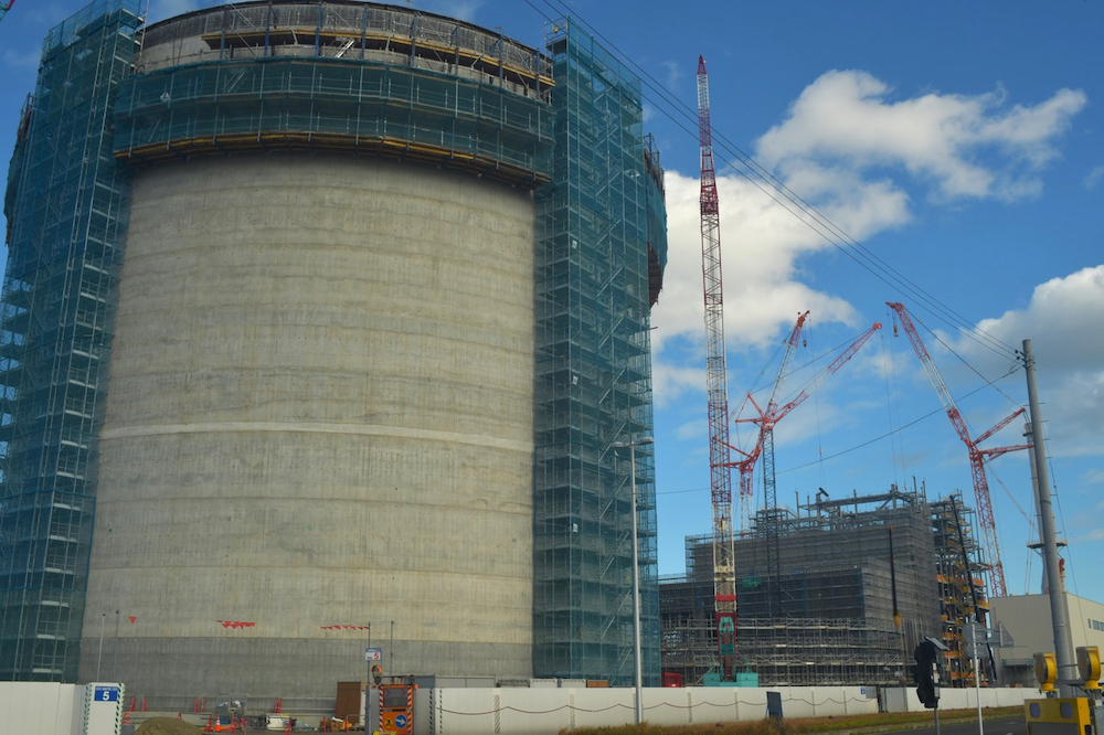 A massive round concrete structure is partly cloaked in green scaffolding. Behind it, cranes tower over a large building under construction.