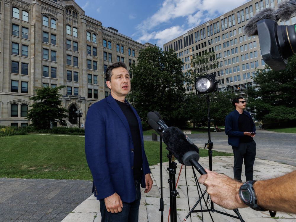 A light-skinned man with short brown hair, wearing a blue jacket over a black shirt, stands in the outdoor courtyard of a building complex and speaks into a microphone being held toward him.