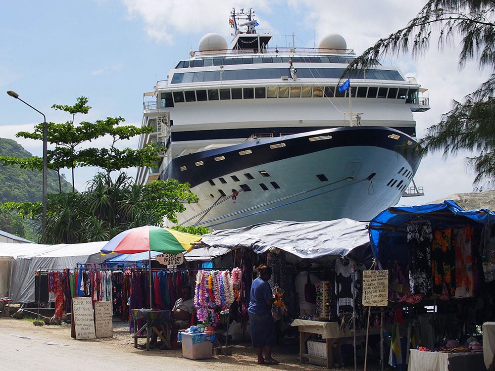 A row of vendors stands are along a dirt road, with the giant bow of a cruise ship looming over them.