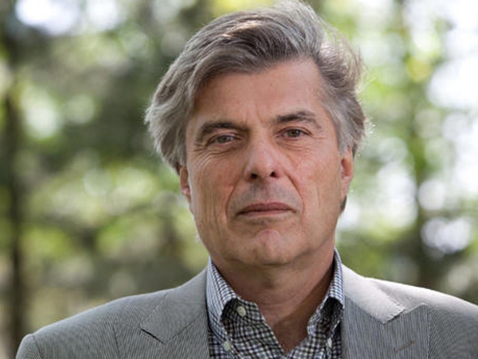 A close-up of a light-skinned man with short grey hair, wearing a grey jacket over a checkered shirt.
