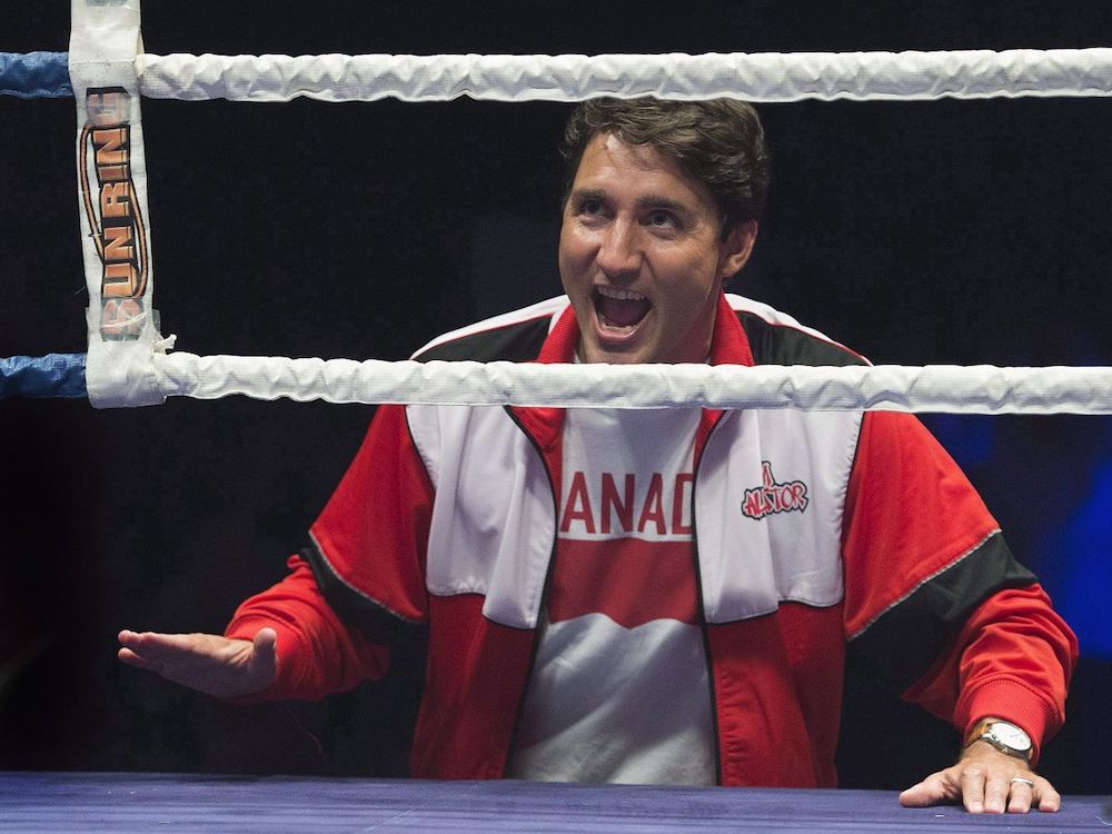 Justin Trudeau, wearing red and white sporty clothing, stands just outside the ropes of a boxing ring, with one hand on the ring floor. His mouth is open and smiling.