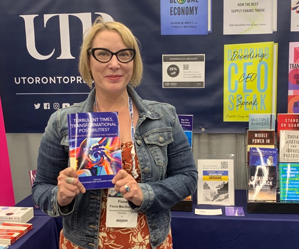A woman with medium-light skin tone, dark rimmed glasses and a blond bob smiles at the camera and holds up a book.