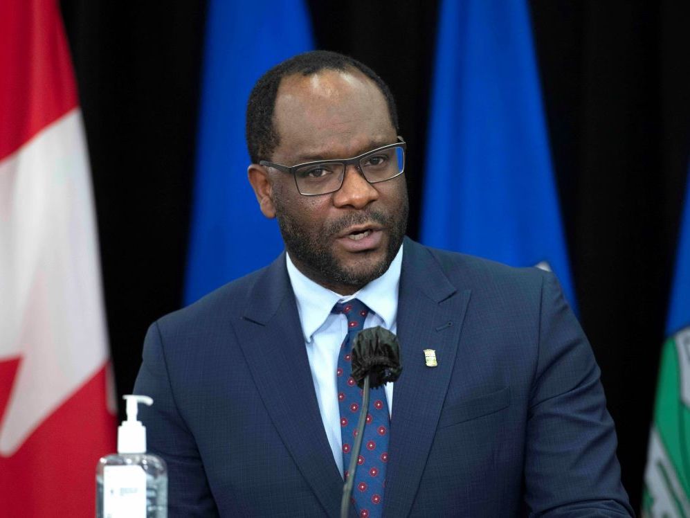 Kaycee Madu is a middle-aged Black man wearing glasses and a navy suit. He stands at a podium and speaks into a microphone. To his right is a bottle of hand sanitizer. In the background are the Alberta and Canada flags.