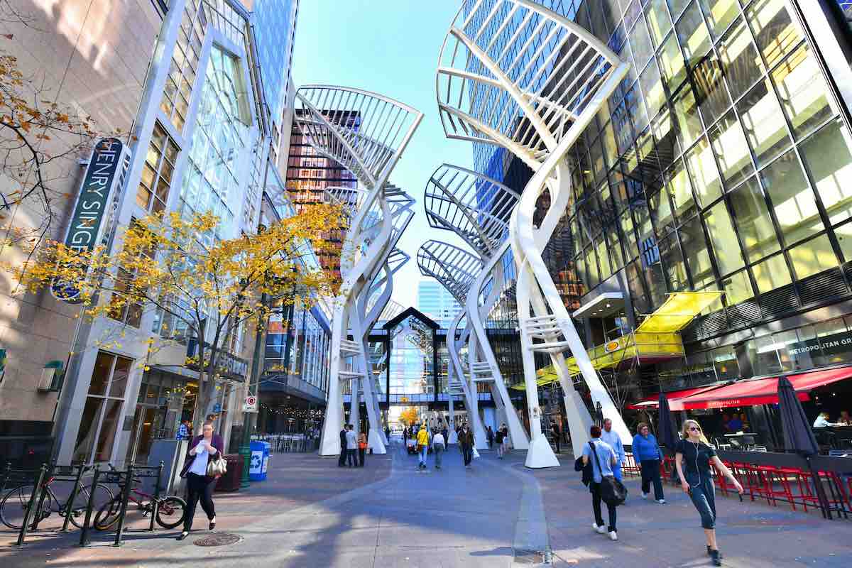 Pedestrians walk along a tiled concrete plaza flanked by tall fluted white public art structures flanked by contemporary highrises for business.