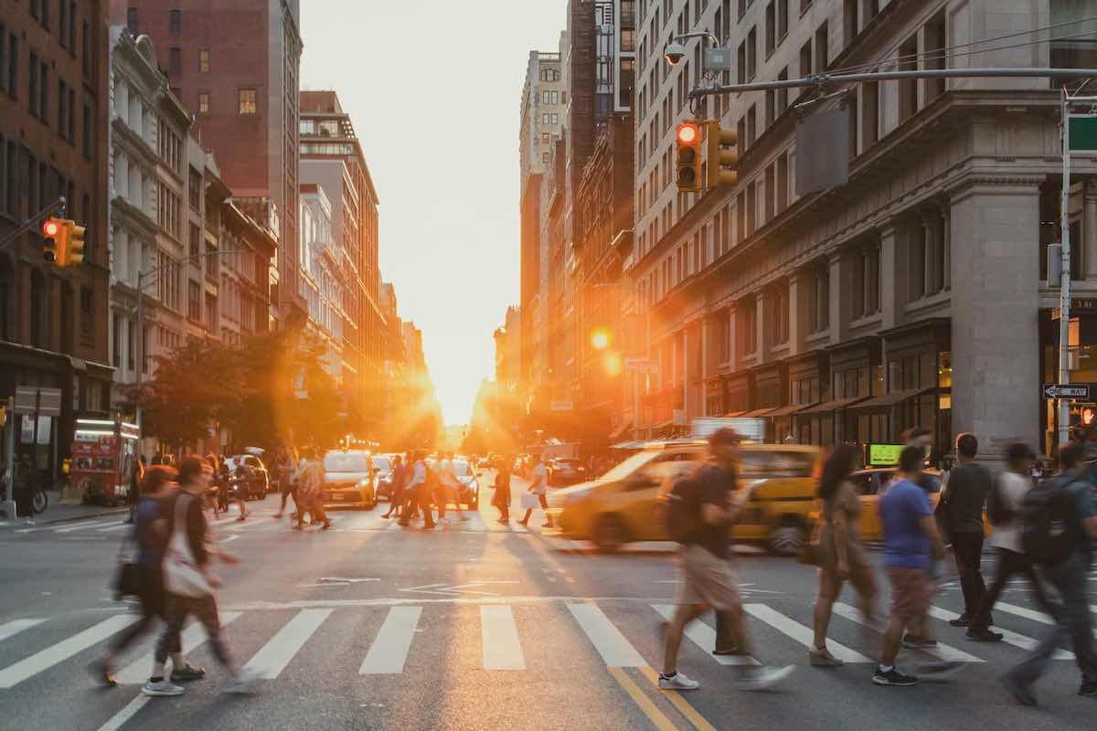 A street shot of pedestrians crossing the street at a crosswalk in New York City. The sun is on the horizon and the pedestrians are flanked by multi-storey brick buildings and the busy traffic of yellow taxis.