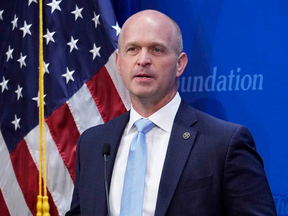 A man with light skin, short buzzed greying hair and a navy blue suit over a white shirt and light blue tie speaks in front of an American flag and a blue wall that reads 'Heritage Foundation.'