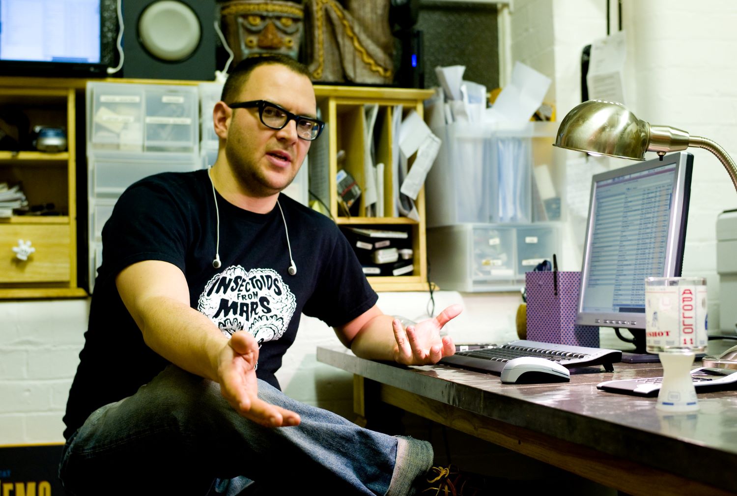 A man with light skin, short dark hair and thick black-framed glasses sits at a desk. He is wearing a black T-shirt with a pair of ear buds dangling from his neck and is gesturing with his hands in mid-speech.