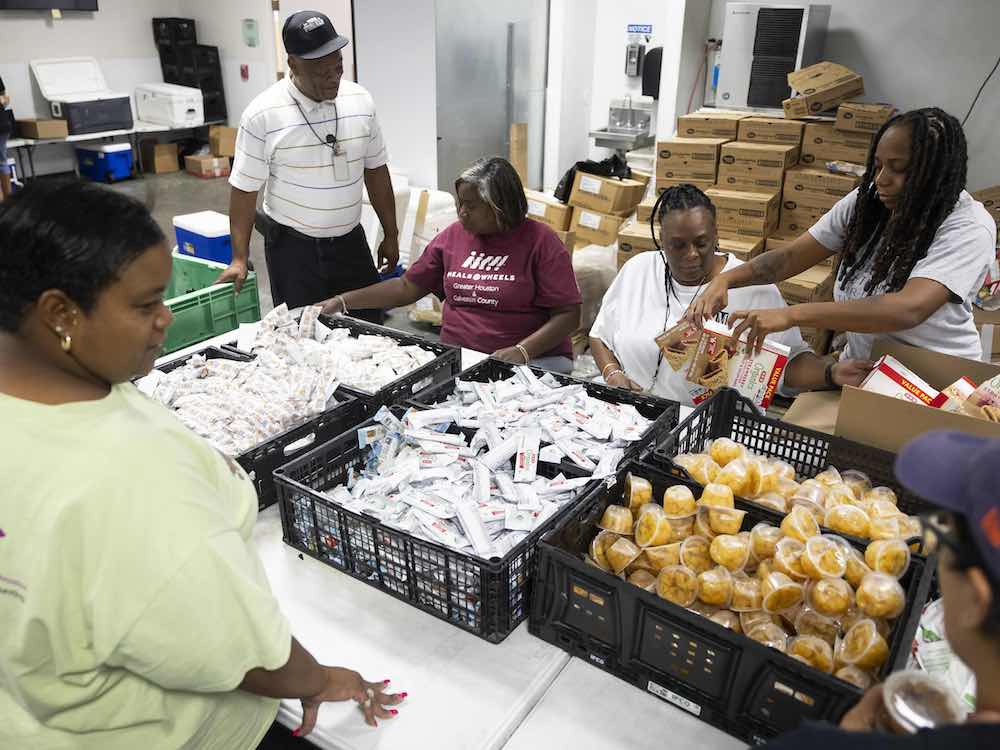 People in summer clothing gather around a table packed with crates of packaged food. They are in a warehouse environment and are packing items as part of a meal delivery program.