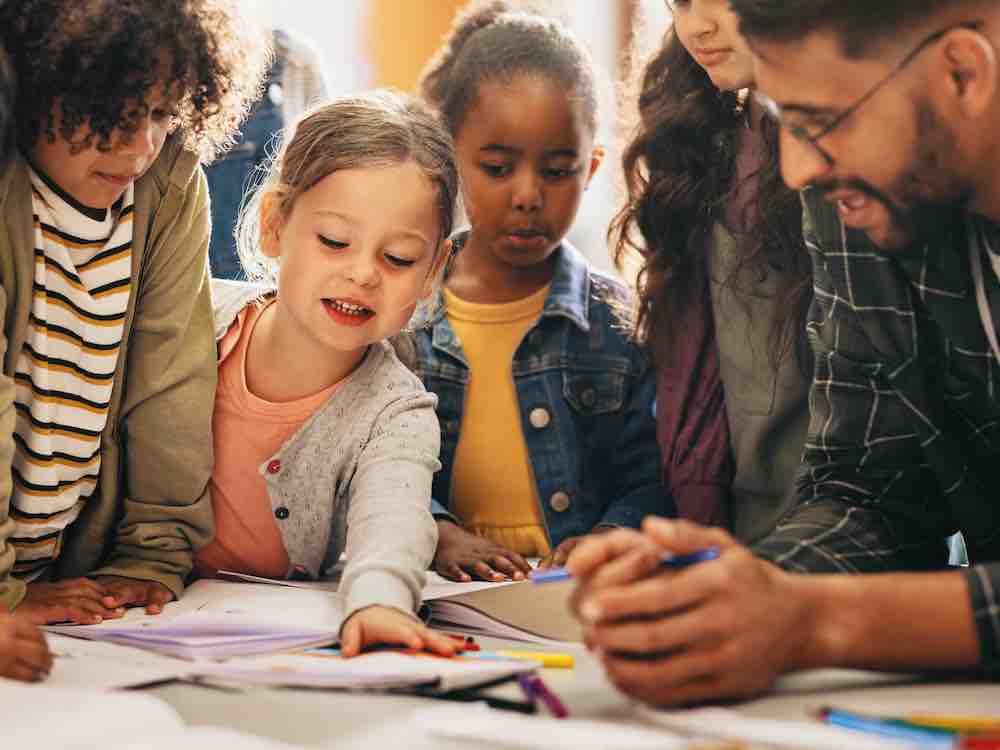 A group of young, racially diverse elementary school students are standing around a table covered with small books and markers. A girl with light skin, hair in a ponytail and a grey cardigan over a pink shirt places her hand on a piece of paper in the centre. To the right, a man with medium skin, a beard and glasses wears a plaid shirt and is mid-speech. He leans his arms on the table.