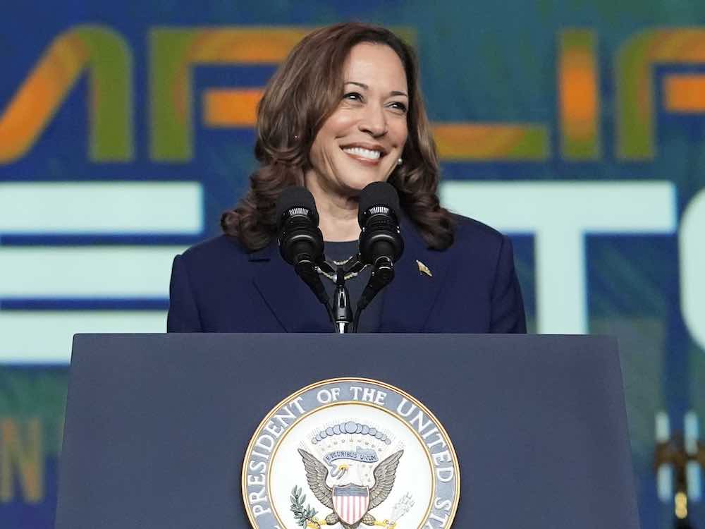 A middle-aged woman with medium skin, shoulder-length brown hair and a navy blazer stands at a podium bearing two black microphones and a crest that reads 'President of the United States.' Behind her is a dark blue background featuring large sans-serif letters in orange, green and white.