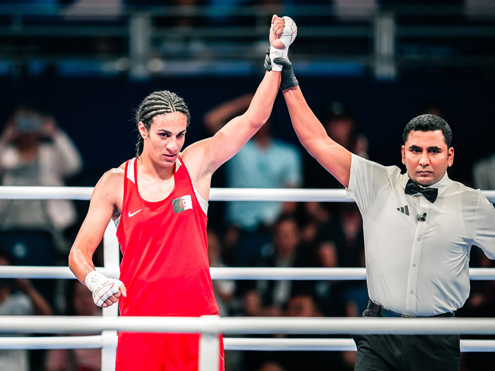 A referee raises Imane Khelif’s fist into the air. She’s wearing red shorts and a red tank top, and her hair is in braids.