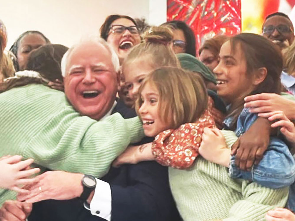 Democratic vice-presidential running mate Tim Walz being hugged and embraced by a group of diverse children. He is buoyantly happy, facing the camera with his mouth wide and smiling and eyes closed.